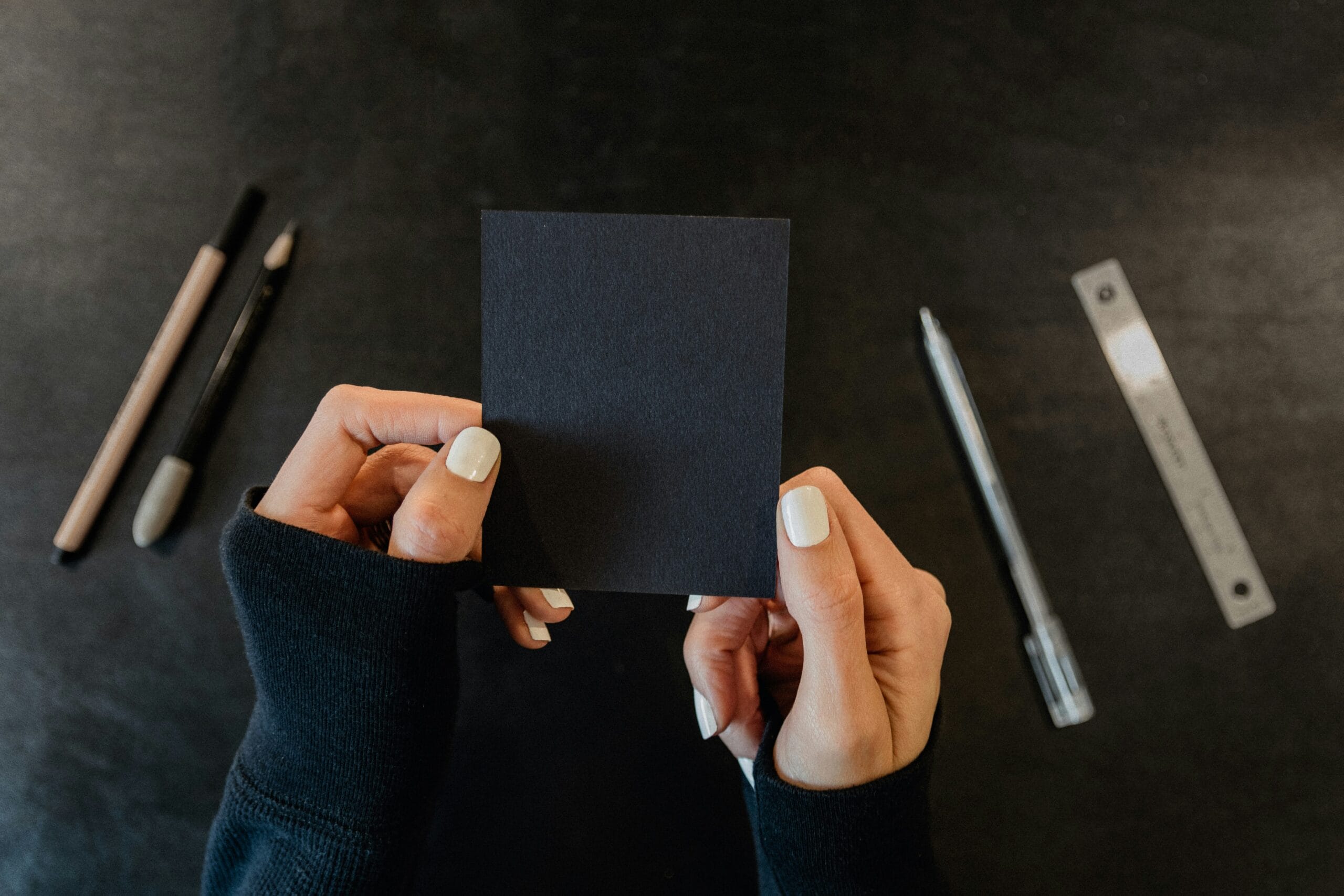 Hands holding a small black card, surrounded by drawing tools and a ruler on a dark surface.