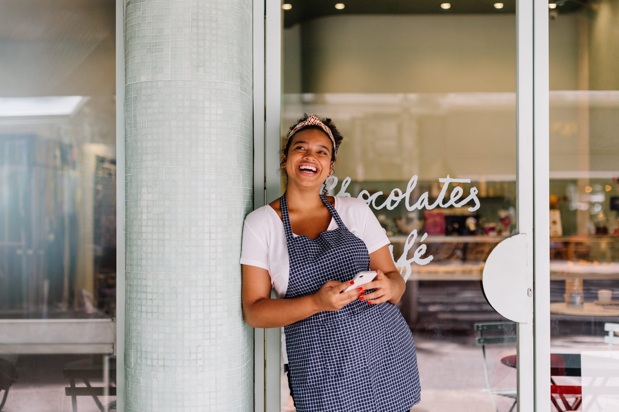 Smiling woman in an apron leans against a column, holding a phone, with a café window displaying "Chocolates."