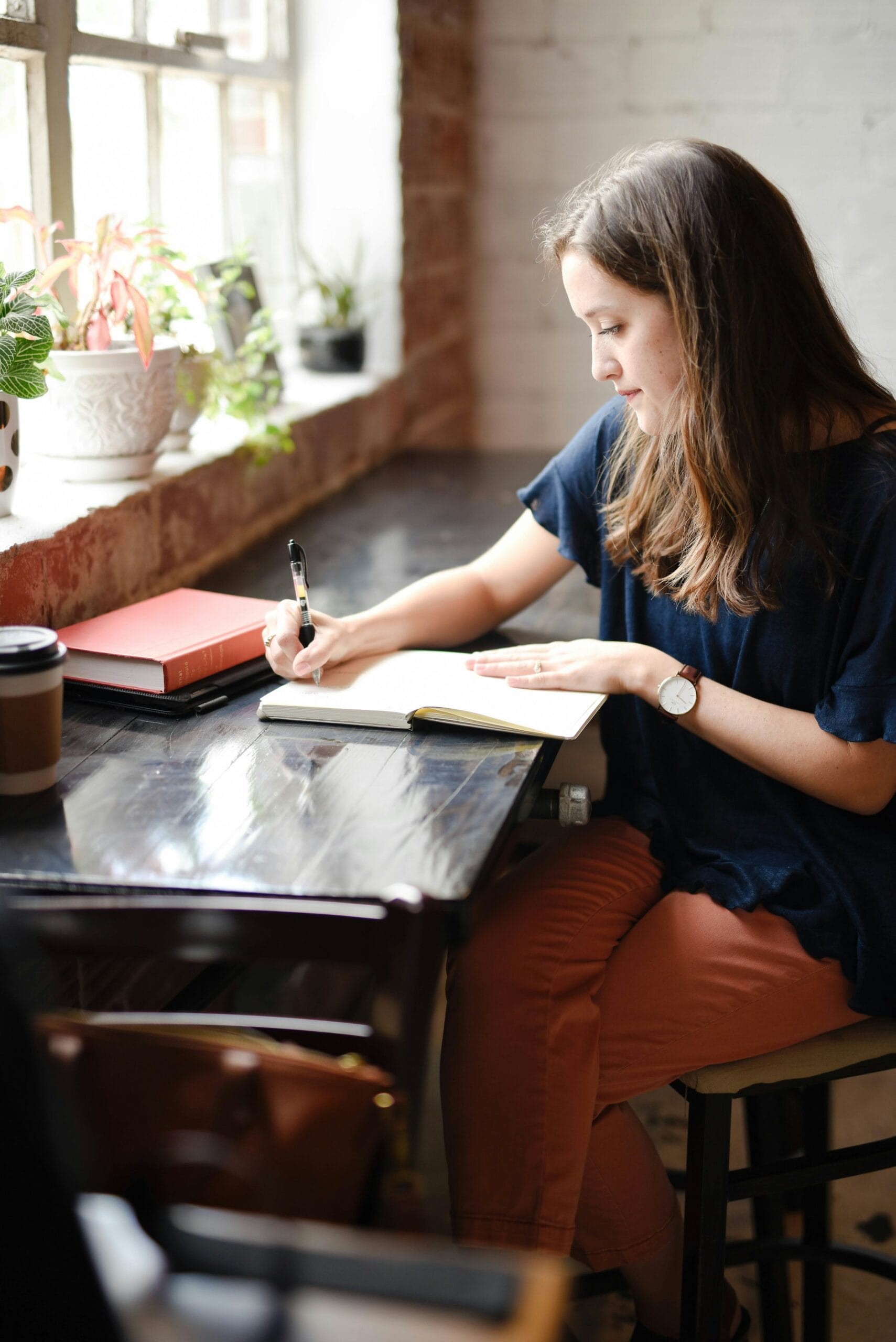 A woman writes in a notebook at a table, surrounded by plants and books, with a coffee cup nearby.