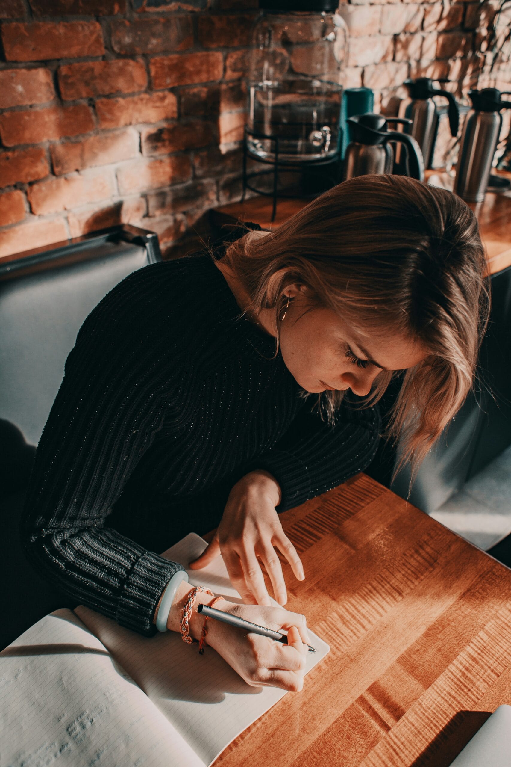 A woman with long hair writes in a notebook at a wooden table, surrounded by a cozy café atmosphere and brick walls.