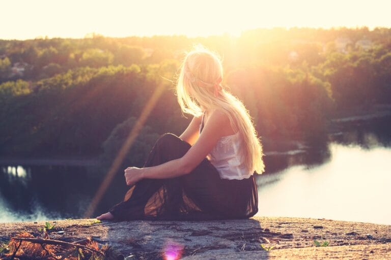 A woman with long hair sits on a rock, gazing at a sunset over a lush landscape and calm water.