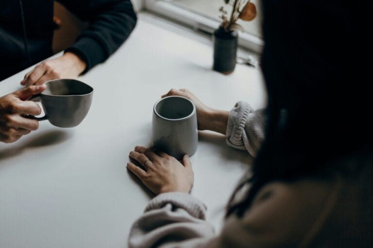 Two hands holding mugs on a table, with a small potted plant in the background and soft, natural lighting.