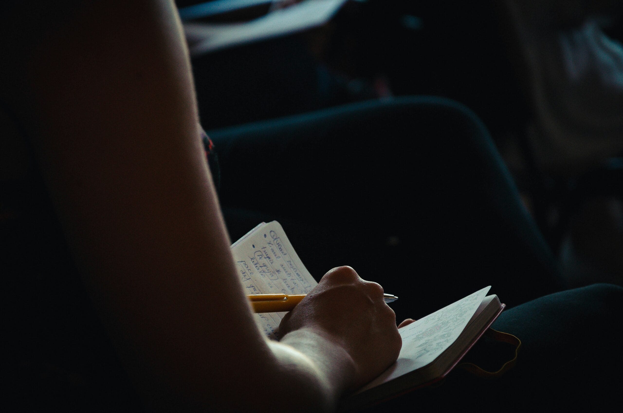 A hand holding a pen writes in a notebook, with a blurred background of seated individuals.
